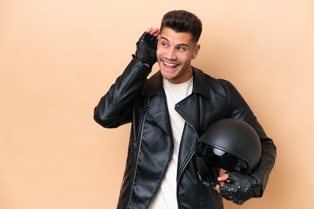 Jeune homme caucasien avec un casque de moto isolé sur fond beige écoutant quelque chose en mettant la main sur l'oreille