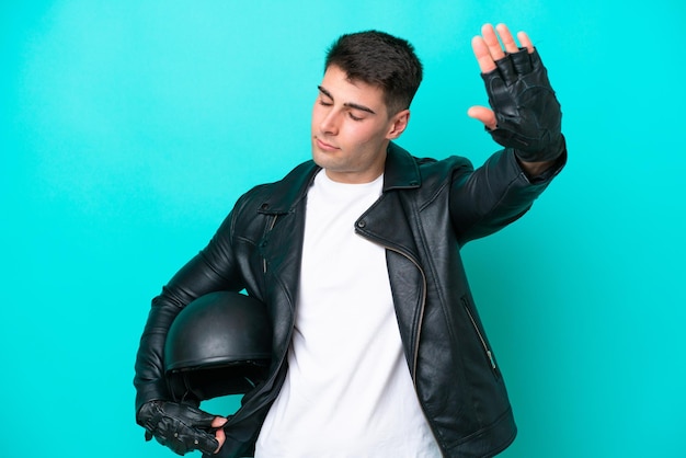 Jeune homme caucasien avec un casque de moto isolé sur fond bleu faisant un geste d'arrêt et déçu