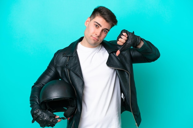 Jeune homme caucasien avec un casque de moto isolé sur fond bleu montrant le pouce vers le bas avec une expression négative