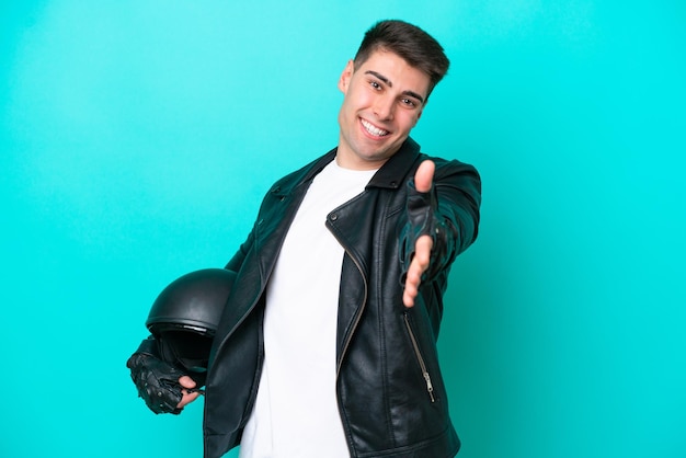 Jeune homme caucasien avec un casque de moto isolé sur fond bleu se serrant la main pour conclure une bonne affaire