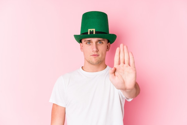 Jeune homme caucasien célébrant la Saint-Patrick debout avec la main tendue montrant le panneau d'arrêt, vous empêchant.