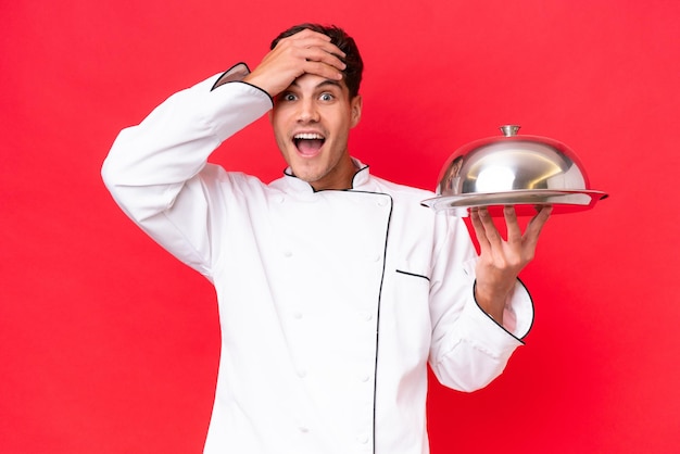 Jeune homme caucasien chef tenant un plateau isolé sur fond rouge avec une expression de surprise