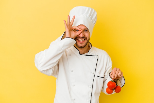 Jeune homme caucasien chef tenant des tomates isolées sur fond jaune excité en gardant le geste ok sur les yeux.