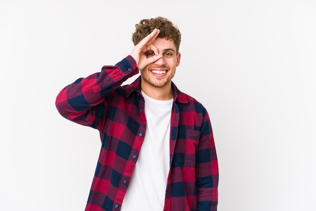 Jeune homme caucasien cheveux bouclés blonds isolé excité en gardant le geste ok sur les yeux.