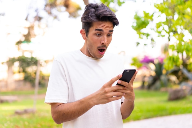 Jeune homme caucasien dans un parc en regardant la caméra tout en utilisant le mobile avec une expression surprise