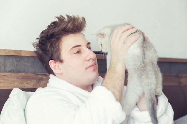 Jeune homme caucasien dans un peignoir tient un chaton blanc. Tôt le matin au lit.