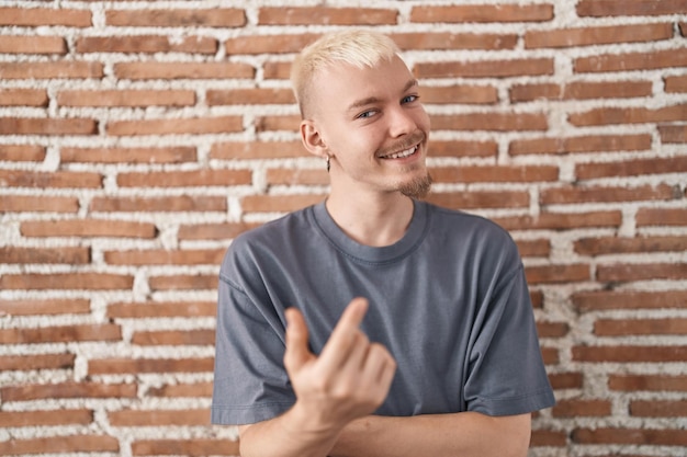 Jeune homme caucasien debout sur un mur de briques faisant signe de venir ici geste avec la main invitant accueillant heureux et souriant