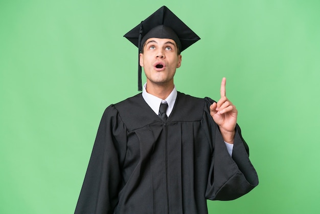 Jeune homme caucasien diplômé universitaire sur fond isolé pointant vers le haut et surpris