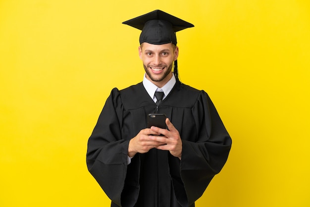 Jeune homme caucasien diplômé universitaire isolé sur fond jaune envoyant un message avec le mobile