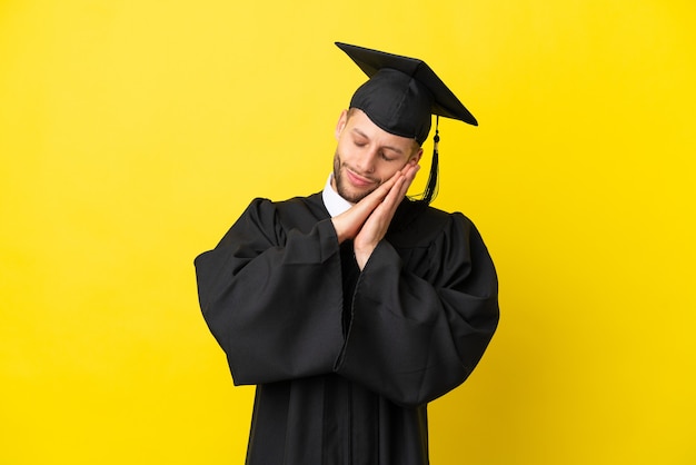 Jeune homme caucasien diplômé universitaire isolé sur fond jaune faisant un geste de sommeil dans une expression adorable