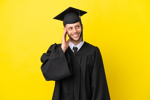 Jeune homme caucasien diplômé universitaire isolé sur fond jaune en pensant à une idée