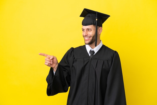Jeune homme caucasien diplômé universitaire isolé sur fond jaune, pointant le doigt sur le côté et présentant un produit