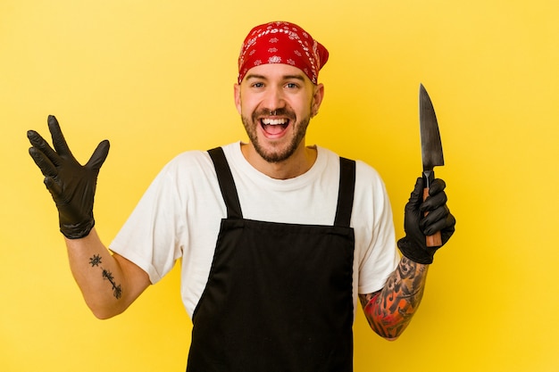 Jeune homme caucasien doseur tatoué tenant un couteau isolé sur un mur jaune recevant une agréable surprise, excité et levant les mains.