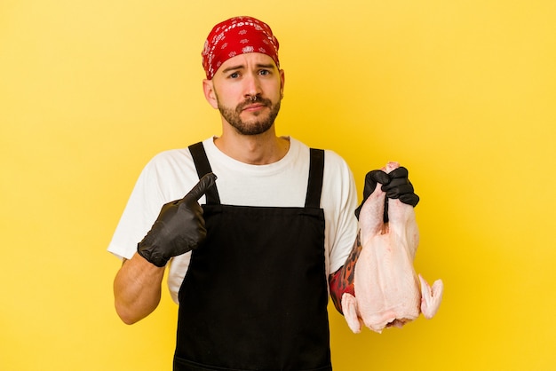 Jeune homme caucasien de doseur tatoué tenant un poulet isolé sur fond jaune pointant avec le doigt sur vous comme si vous invitiez à vous rapprocher.