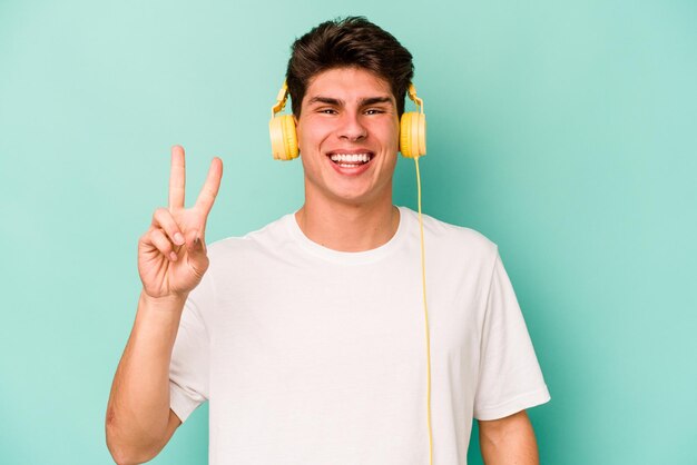 Jeune homme caucasien écoutant de la musique isolé sur fond bleu montrant le numéro deux avec les doigts