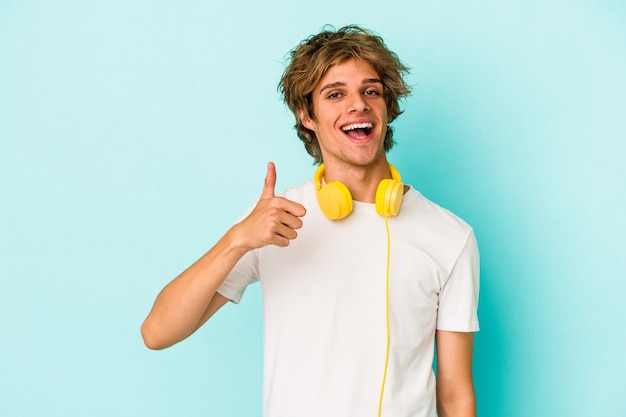 Jeune homme caucasien écoutant de la musique isolé sur fond bleu souriant et levant le pouce vers le haut