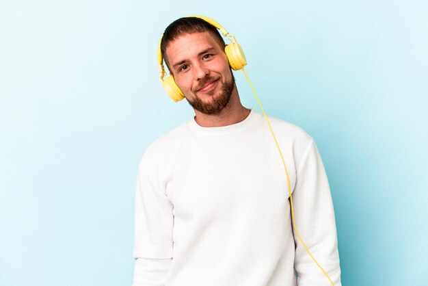 Jeune homme caucasien écoutant de la musique isolée sur fond bleu heureux, souriant et joyeux.