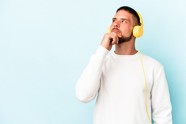 Jeune homme caucasien écoutant de la musique isolée sur fond bleu regardant de côté avec une expression douteuse et sceptique.