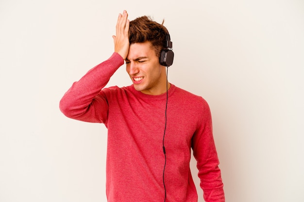 Jeune homme caucasien, écouter de la musique isolée sur un mur blanc en oubliant quelque chose, en frappant le front avec la paume et en fermant les yeux.