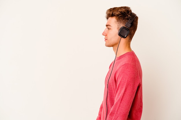 Jeune homme caucasien, écouter de la musique isolée sur un mur blanc regardant à gauche, pose sur le côté.