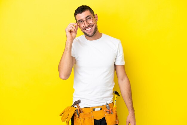 Jeune homme caucasien électricien isolé sur fond jaune avec des lunettes et heureux