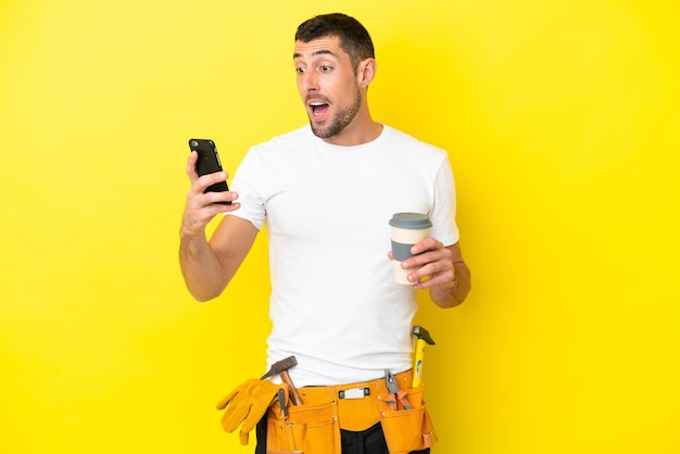 Jeune homme caucasien électricien isolé sur fond jaune tenant du café à emporter et un mobile