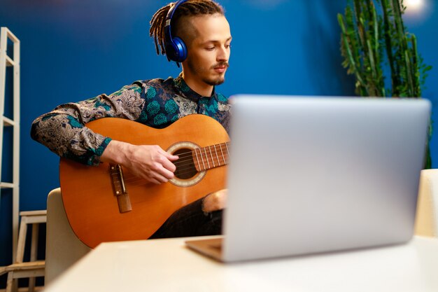 Jeune homme caucasien élégant prenant des cours de guitare en ligne à la maison en utilisant son ordinateur portable