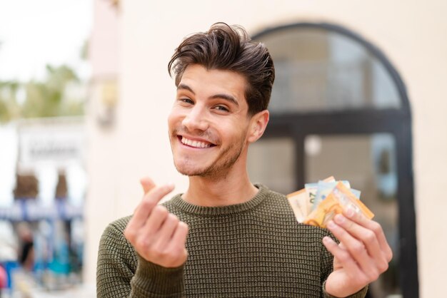 Jeune homme caucasien à l'extérieur prenant beaucoup d'argent avec une expression heureuse