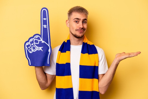 Jeune homme caucasien fan de sport isolé sur fond jaune montrant un espace de copie sur une paume et tenant une autre main sur la taille.