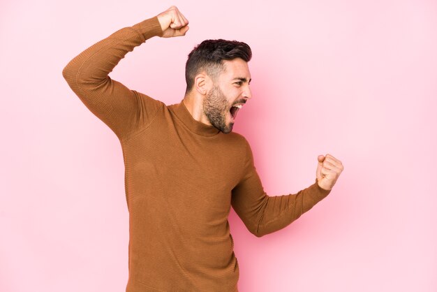 Jeune homme caucasien sur fond rose isolé levant le poing après une victoire, concept gagnant.