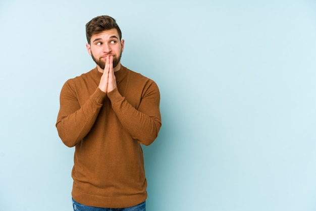 Jeune homme caucasien isolé sur bleu priant, montrant la dévotion, la personne religieuse à la recherche d'inspiration divine.