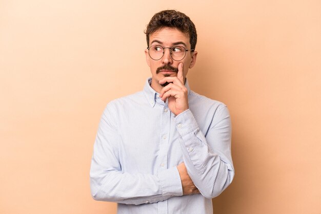 Jeune homme caucasien isolé sur fond beige contemplant, planifiant une stratégie, réfléchissant à la voie d'une entreprise.