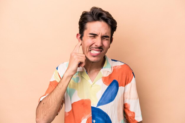 Jeune homme caucasien isolé sur fond beige couvrant les oreilles avec les mains