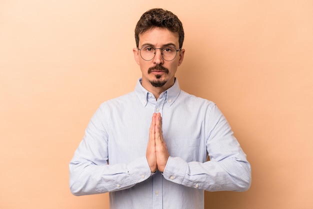 Jeune homme caucasien isolé sur fond beige priant, montrant la dévotion, personne religieuse à la recherche d'inspiration divine.