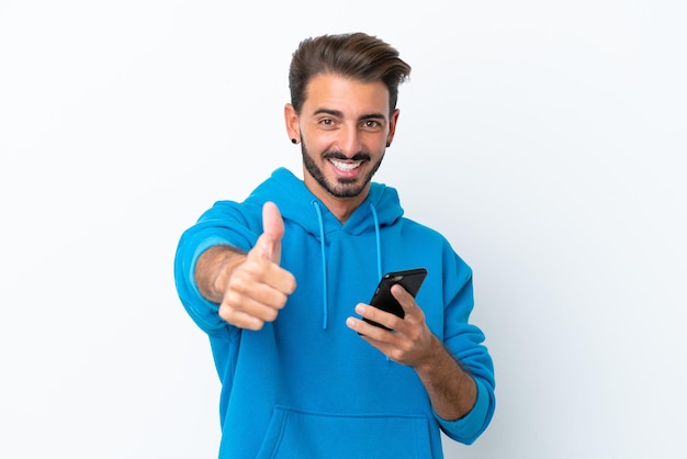 Jeune homme caucasien isolé sur fond blanc à l'aide de téléphone portable tout en faisant les pouces vers le haut