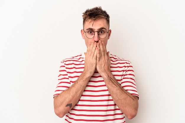 Jeune homme caucasien isolé sur fond blanc choqué, couvrant la bouche avec les mains, impatient de découvrir quelque chose de nouveau.