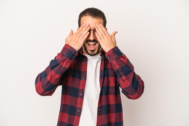 Jeune homme caucasien isolé sur fond blanc couvre les yeux avec les mains sourit largement en attente d'une surprise