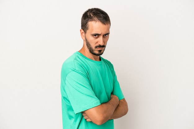 Jeune homme caucasien isolé sur fond blanc, fronçant les sourcils de mécontentement, garde les bras croisés.