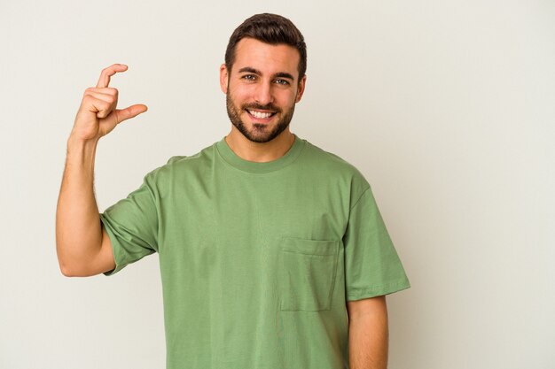 Jeune homme caucasien isolé sur fond blanc tenant quelque chose de peu avec l'index, souriant et confiant.