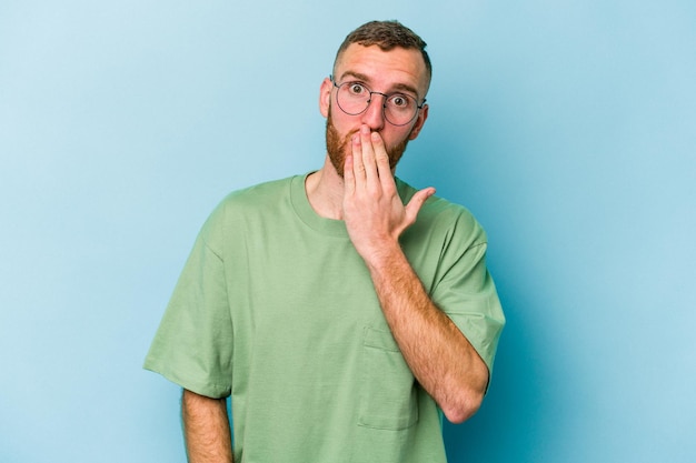 Jeune homme caucasien isolé sur fond bleu choqué, couvrant la bouche avec les mains, soucieux de découvrir quelque chose de nouveau.