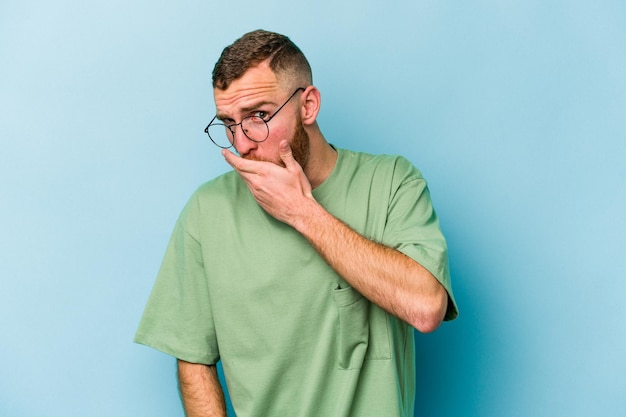 Jeune homme caucasien isolé sur fond bleu couvrant la bouche avec les mains semblant inquiet.