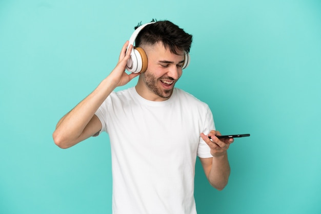 Jeune homme caucasien isolé sur fond bleu, écouter de la musique avec un mobile et chanter