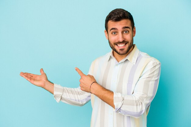 Jeune homme caucasien isolé sur fond bleu excité tenant un espace de copie sur la paume.