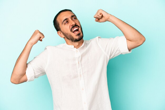 Jeune homme caucasien isolé sur fond bleu levant le poing après une victoire, concept gagnant.