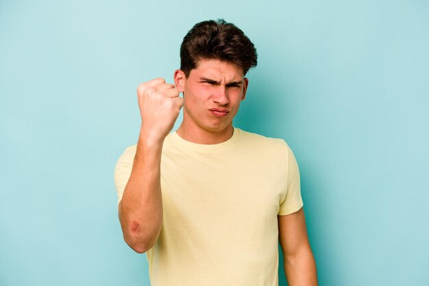 Jeune homme caucasien isolé sur fond bleu montrant le poing à la caméra expression faciale agressive