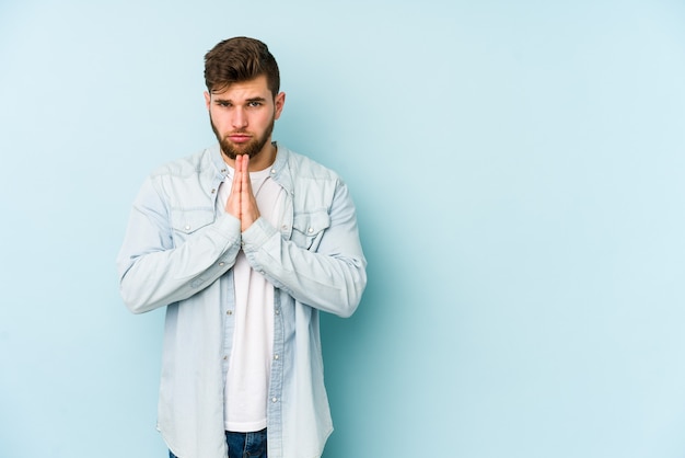 Jeune homme caucasien isolé sur fond bleu priant, montrant la dévotion, personne religieuse à la recherche d'inspiration divine.