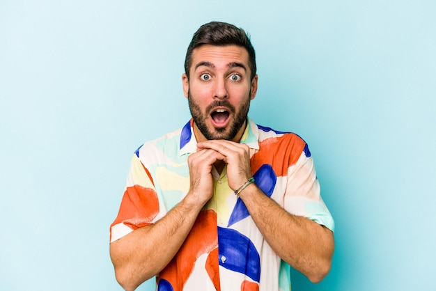 Jeune homme caucasien isolé sur fond bleu priant pour la chance étonné et ouvrant la bouche regardant vers l'avant