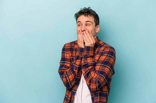 Jeune homme caucasien isolé sur fond bleu riant de quelque chose, couvrant la bouche avec les mains.