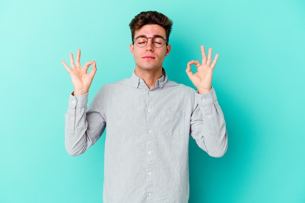 Jeune homme caucasien isolé sur fond bleu se détend après une dure journée de travail, elle effectue du yoga.