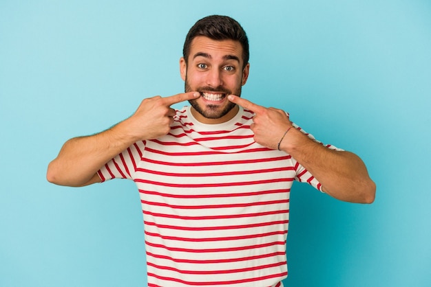 Jeune homme caucasien isolé sur fond bleu sourit, pointant du doigt la bouche.